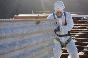 A worker in a white protective suit, helmet, and respirator removing insulation material from a structure with wooden beams in the background.