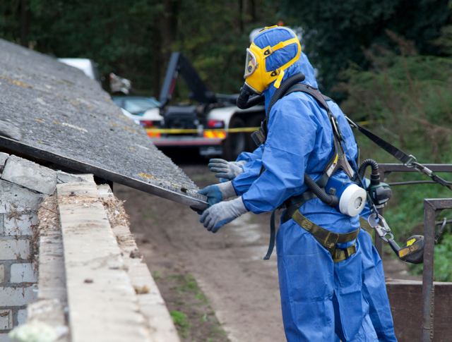 A worker in a blue protective suit with a yellow and blue respirator removing an asbestos roof section.