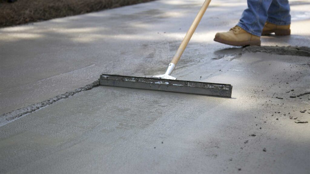 A worker smoothing fresh concrete on a walkway with a long-handled tool.