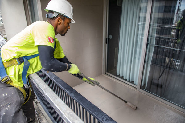 A worker in a safety harness and hard hat using a tool to clean a balcony near a glass door.