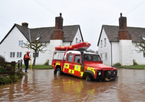 Two firefighters in orange gear working near a red rescue vehicle in floodwaters in front of a house.