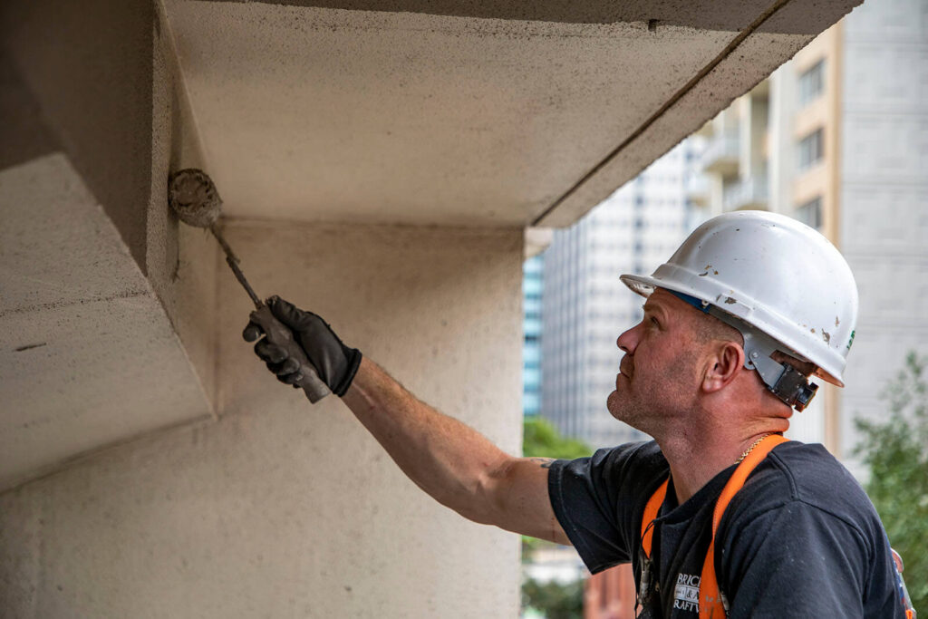 A worker in a helmet and safety vest applying concrete repair material under a balcony.