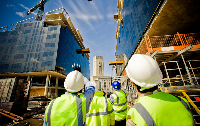 A group of construction workers in safety vests and hard hats walking on a site with cranes and buildings in the background.