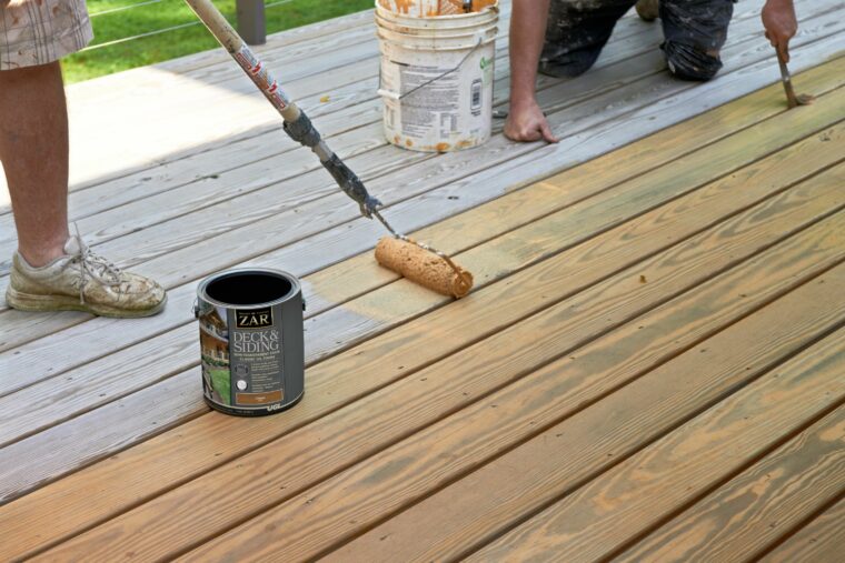 Two workers using a brush and roller to paint a wooden deck with a ZAR paint can.